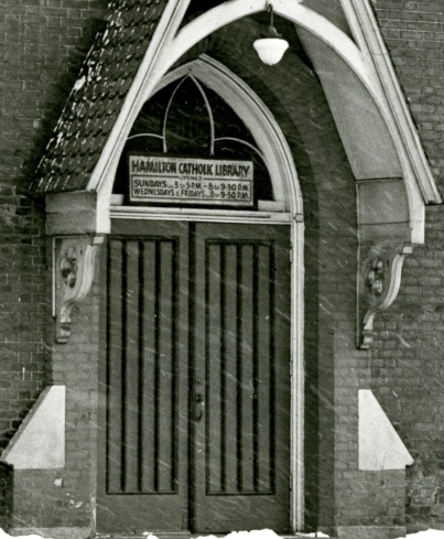 Black and white photograph of the front of a church with a sign over the door for "Hamilton Catholic Library"